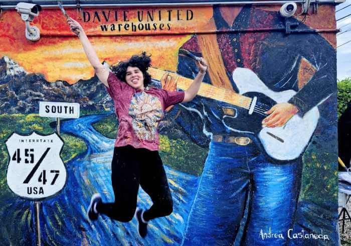 Vibrant outdoor mural featuring a guitarist, highway sign, and energetic motion, painted by artist Andrea Castañeda