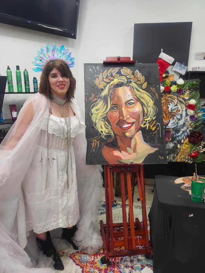Artist Andrea Castañeda standing beside her live portrait painting on an easel during a painting performance, showcasing expressive brushwork and contemporary figurative art.