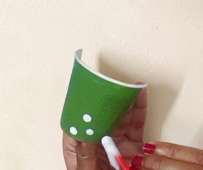 Hand painting small white dots on a green paper cup to create decorative elements for DIY cardboard wall decor